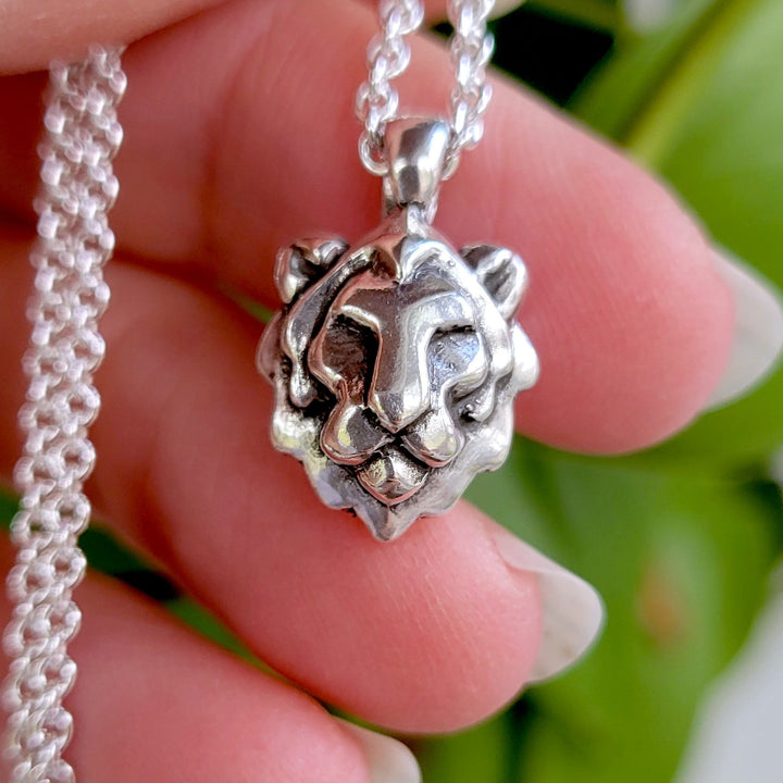Silver necklace with a lion head pendant held by a hand against a green leafy background